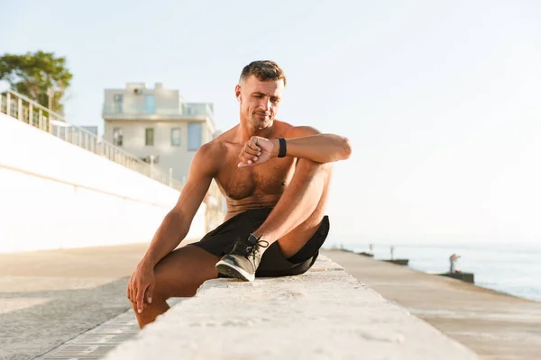 해변에 앉아있는 스마트 시계를 이어폰 잘생긴 Shirtless 스포츠맨 — 스톡 사진