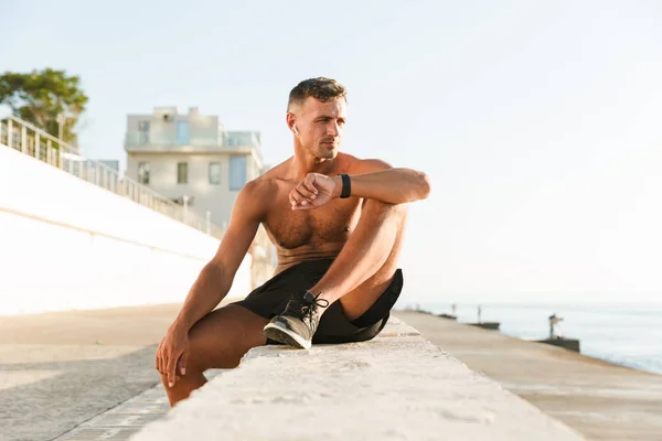 해변에 앉아있는 스마트 시계를 이어폰 자신감 잘생긴 Shirtless 스포츠맨 — 스톡 사진