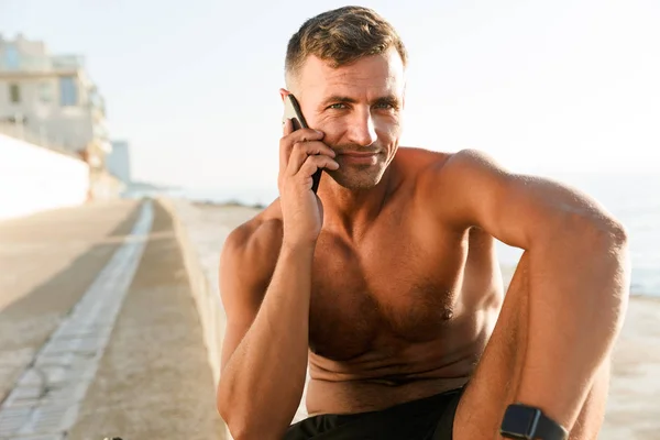 Image of handsome strong mature sportsman talking by mobile phone on the beach outdoors.