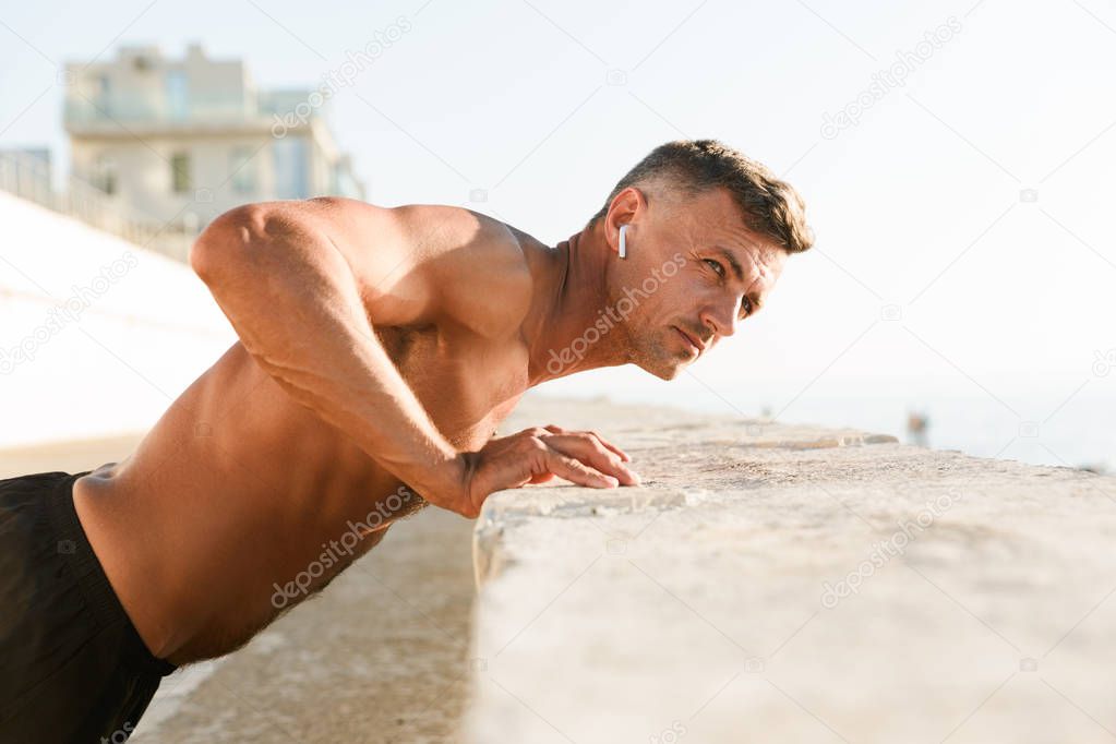 Image of handsome strong mature sportsman make sport exercises on the beach outdoors listening music with earphones.