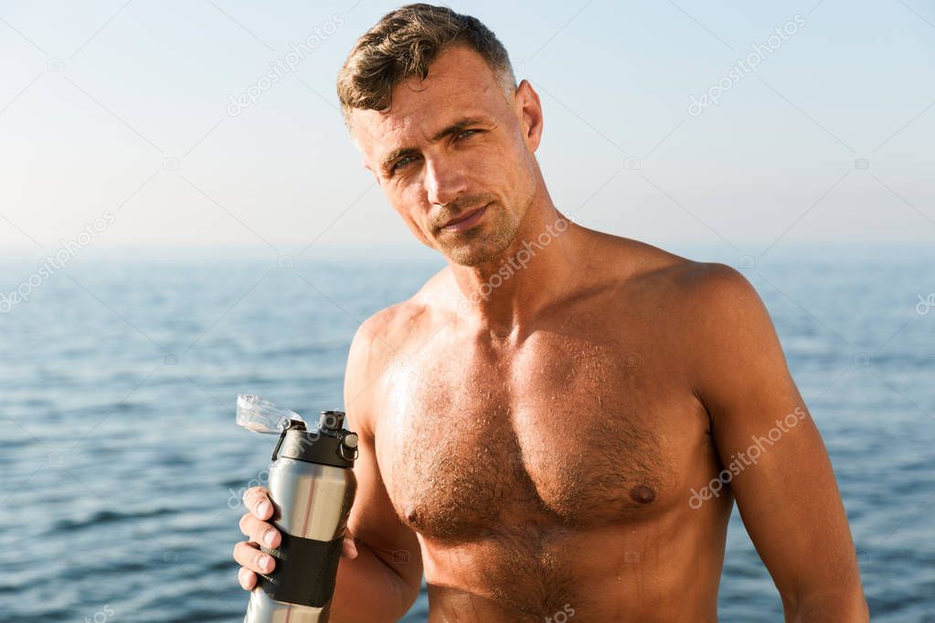 Smiling handsome shirtless sportsman holding water bottle while standing at the beach