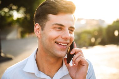 Cep telefonu açık havada üzerinde konuşurken gömlek gülümseyen genç bir adamın yakın çekim