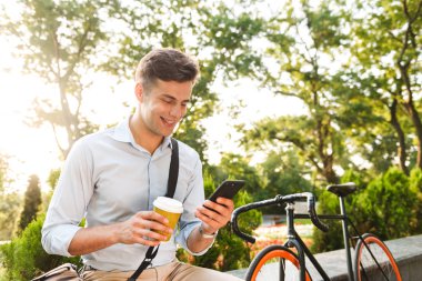 Kahve ve bisiklet açık havada bir bardak ile otururken telefon kullanarak gömlek gülümseyen genç bir adam portresi giymiş