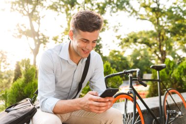 Genç gülümseyen bir adam portresi ile bisiklet açık havada otururken telefon kullanarak gömlek giymiş
