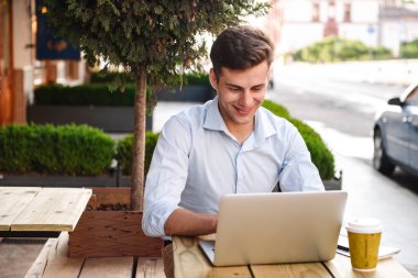 Şık delikanlı bir kafe açık havada otururken dizüstü bilgisayarda çalışarak gülümseyen