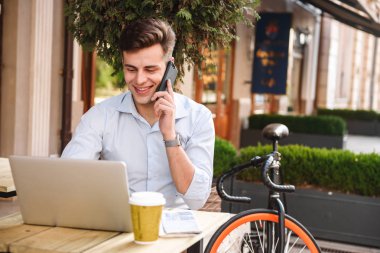 Şık delikanlı ile dizüstü bilgisayar açık havada bisiklet ile bir kafede otururken cep telefonu üzerinde konuşurken gömlek gülümseyen