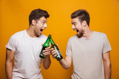 Portre bir iki genç erkek arkadaşlarım bira şişeleri ile toasting sarı arka plan üzerinde izole heyecanlı