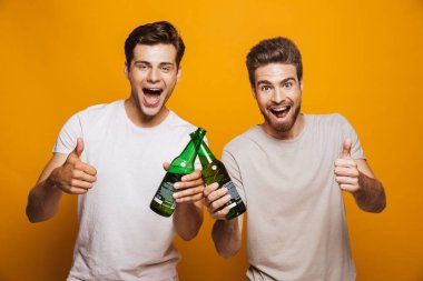 İki mutlu genç adam portresi en başparmak sarı arka plan üzerinde izole gösterilmesini bira şişeleri ile toasting yakın arkadaşlarım