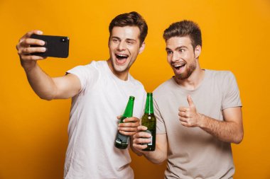 Bir iki neşeli genç adam portresi en bira şişesi holding ve sarı arka plan üzerinde izole bağırarak süre bir selfie alarak yakın arkadaşlarım