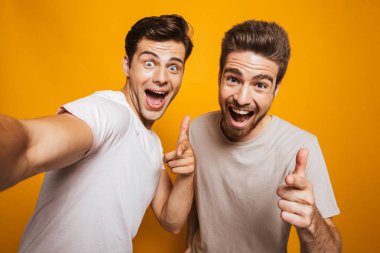 Arkadaşlarım bir selfie alarak kameraya parmakları işaret sarı arka plan üzerinde izole iki mutlu genç adam portresi