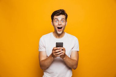 Cep telefonu izole sarı arka plan üzerinde tutan bir şok genç adam portresi kutluyor