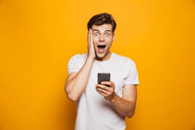 Cep telefonu izole sarı arka plan üzerinde tutarak şaşırmış gibi bir genç adam portresi kutluyor