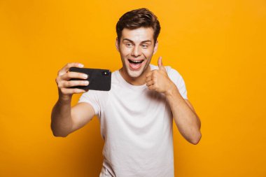 Başparmak gösterilen sarı arka plan üzerinde izole bir selfie alarak neşeli genç bir adam portresi