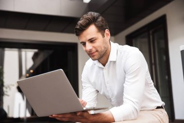 Açık havada otururken dizüstü bilgisayar üzerinde çalışan bir kendine güvenen genç resmi giyimli adam portresi