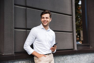 Cep telefonu açık havada tutan bir duvara yaslanmış bir gülümseyen genç resmi giyimli adam portresi