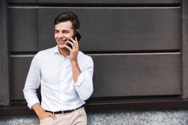 Açık havada cep telefonda konuşurken bir duvara yaslanmış bir gülümseyen genç resmi giyimli adam portresi