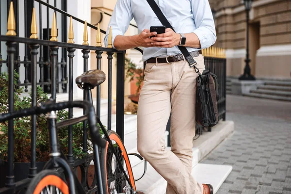 若いキャリング バッグ街で自転車と一緒に立っていると 携帯電話を保持しているシャツに身を包んだスタイリッシュな男の画像をトリミング — ストック写真