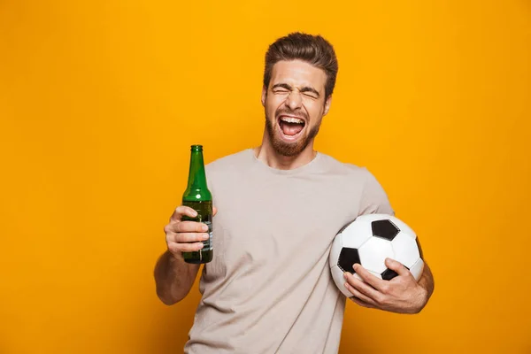 黄色の背景に分離したビール瓶とサッカー ボールを持って幸せな若い男の肖像 — ストック写真