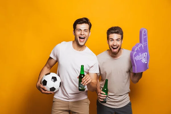 Portrait of a two cheerful young men best friends with soccer ball holding beer bottles and shouting isolated over yellow background
