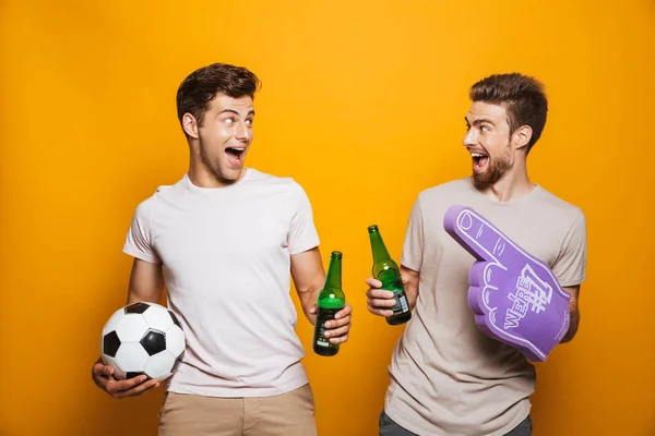 Portrait of a two excited young men best friends with soccer ball toasting with beer bottles and shouting isolated over yellow background