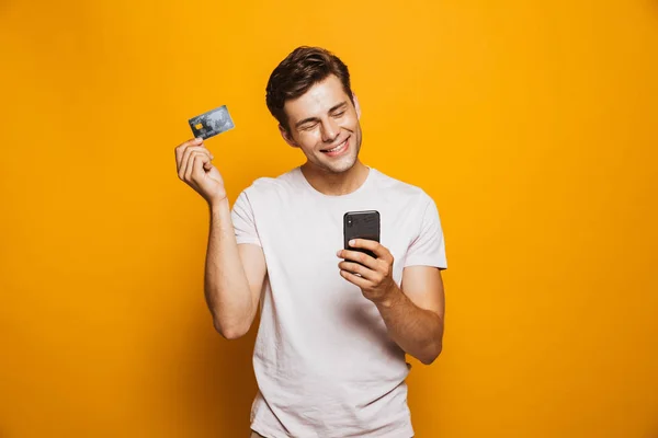 一个满意的年轻人手持手机和显示在黄色背景下隔离信用卡的肖像 — 图库照片