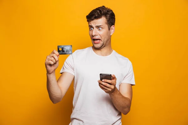 一个沮丧的年轻人手持手机和显示在黄色背景下隔离信用卡的肖像 — 图库照片
