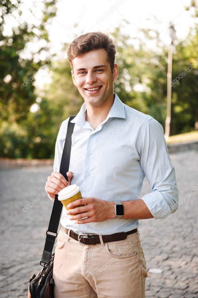 Smiling young man in shirt carrying bag walking at the city park with cup of coffee