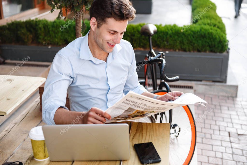 Happy young stylish man in shirt reading newspaper while sitting with laptop computer at a cafe outdoors with bicycle