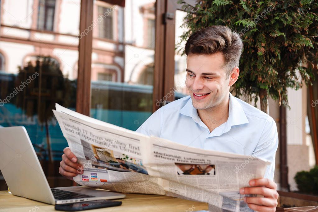 Smiling young stylish man in shirt reading newspaper while sitting with laptop computer at a cafe outdoors