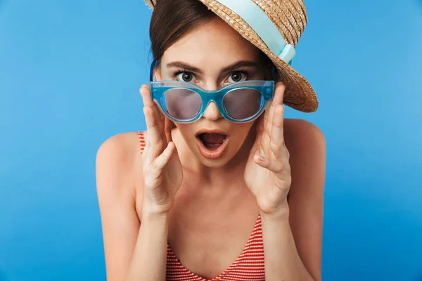 一个惊讶的年轻女孩穿着泳装的肖像戴着太阳镜和夏天的帽子摆在蓝色背景下孤立 — 图库照片