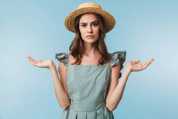 关闭在礼服和夏天帽子的一个困惑的女人的肖像耸耸肩肩膀孤立的蓝色背景 — 图库照片