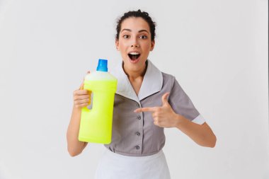 Beyaz arka plan üzerinde izole deterjan tutan üniforması giymiş bir heyecanlı genç hizmetçi portresi