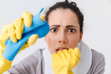 Beyaz arka plan üzerinde izole bir sünger kullanarak üniforma ve lastik eldiven giymiş korkmuş genç hizmetçi portresi