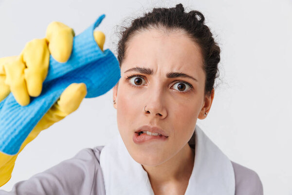 Portrait of a confused young housemaid dressed in uniform and rubber gloves using a sponge isolated over white background