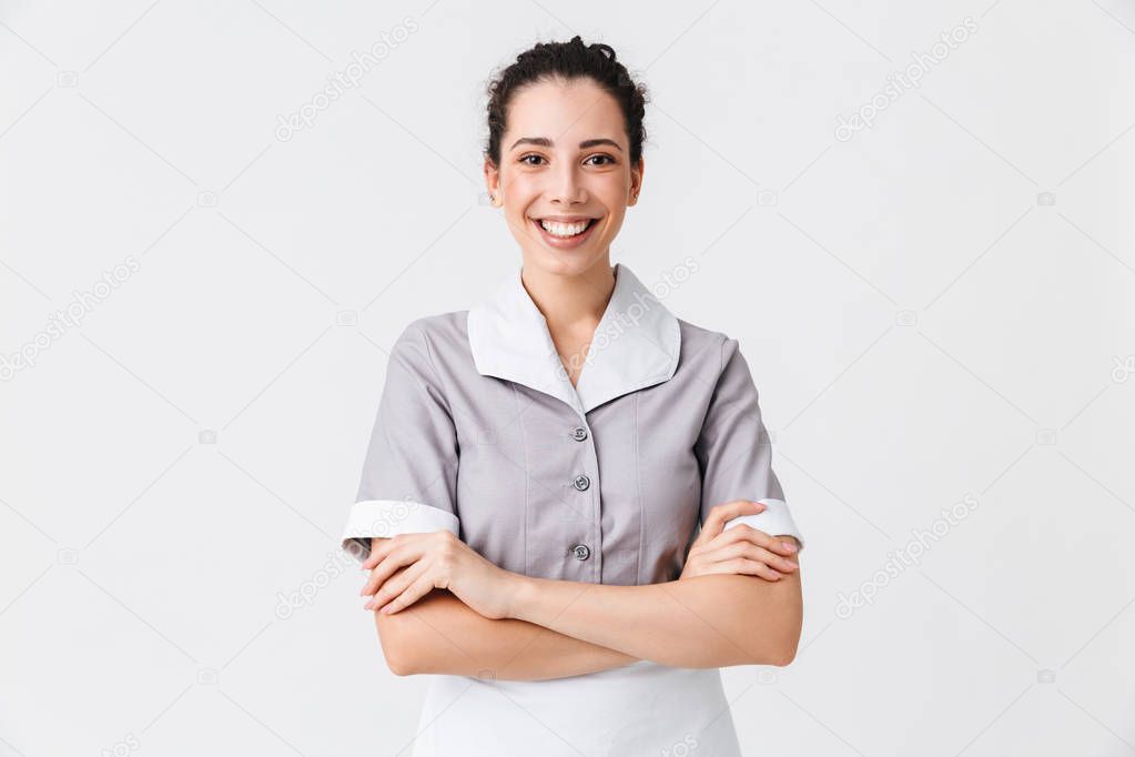 Portrait of a cheerful young housemaid dressed in uniform standing with arms folded isolated over white background