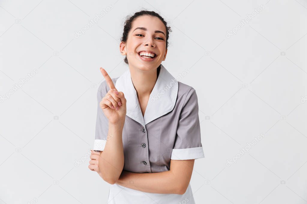 Portrait of a cheerful young housemaid dressed in uniform pointing finger up isolated over white background