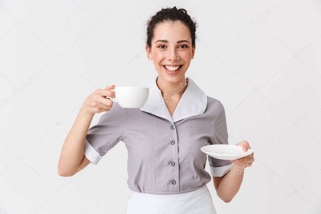 Portrait of a smiling young housemaid dressed in uniform holding cup with a saucer isolated over white background