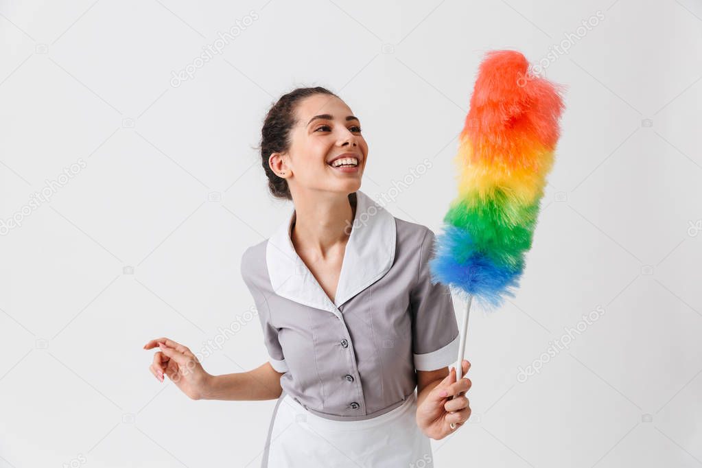 Portrait of a pretty young housemaid dressed in uniform holding a duster isolated over white background