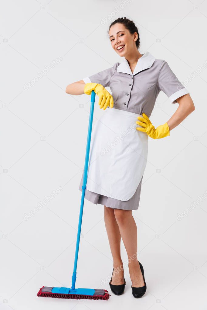 Full length portrait of a happy young housemaid dressed in uniform holding a mop isolated over white background