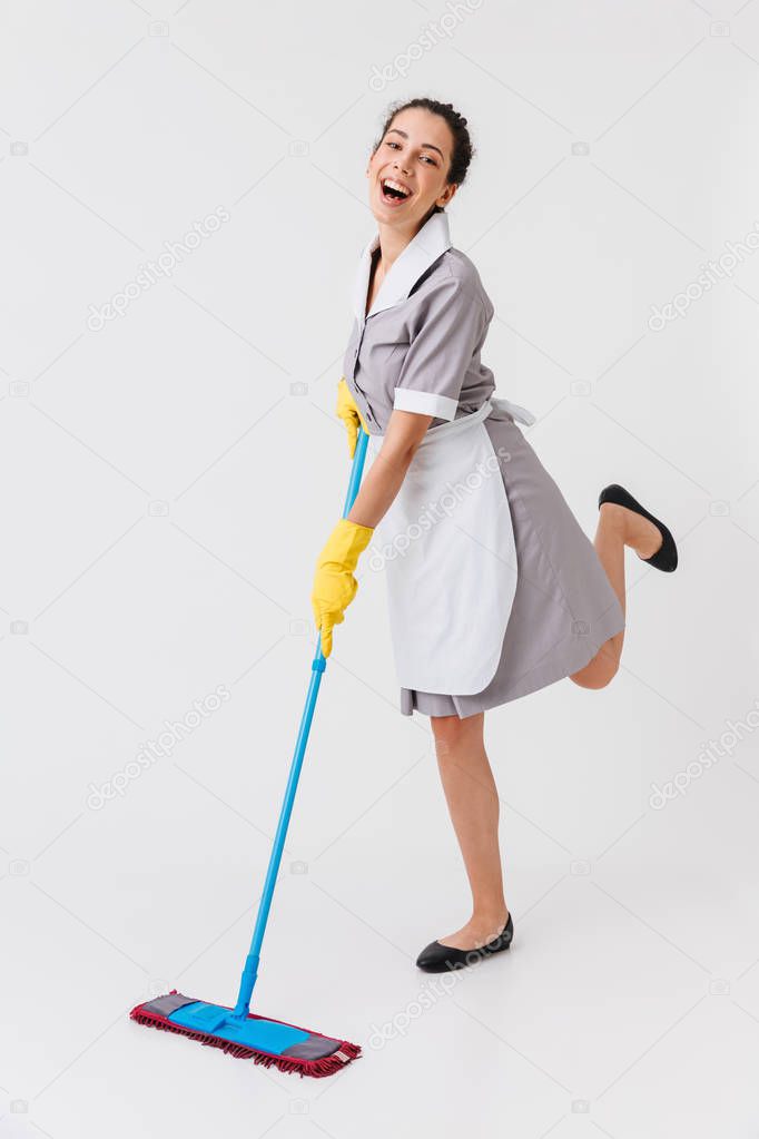 Full length portrait of a joyful young housemaid dressed in uniform using a mop isolated over white background