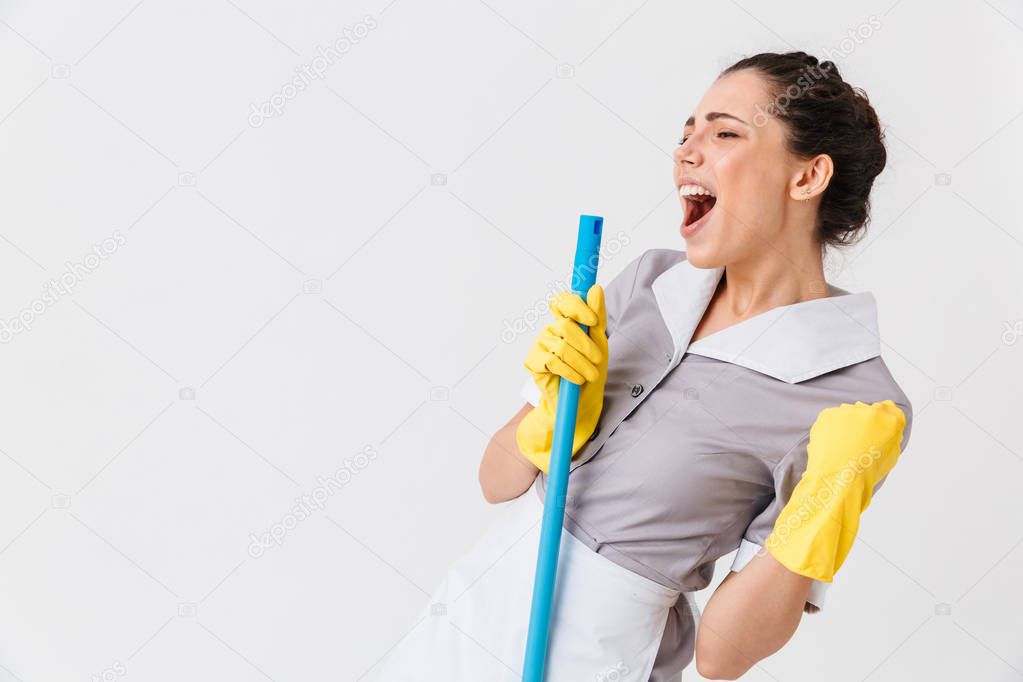 Portrait of an excited young housemaid dressed in uniform using a mop isolated over white background