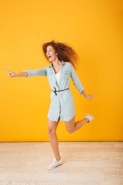 Gülümseyen ve işaret eden 20'li yaşlarda bir kenara boşaltmak izole sarı arka plan üzerinde parmağını. şaşırmış kıvırcık kadın portresi uzunluğu tam