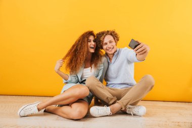 Neşeli erkek ve kız 20'li yaşlarda fotoğrafı katta birlikte ve alma selfie sarı arka plan üzerinde izole smartphone üzerinde oturan