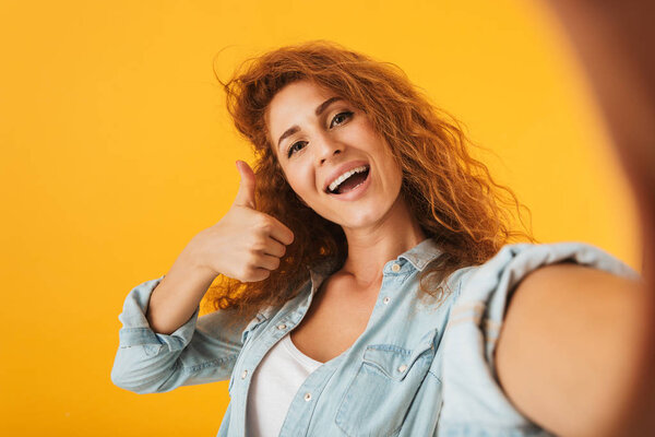 Photo of curly attractive woman 20s smiling and showing thumb up while taking selfie photo isolated over yellow background