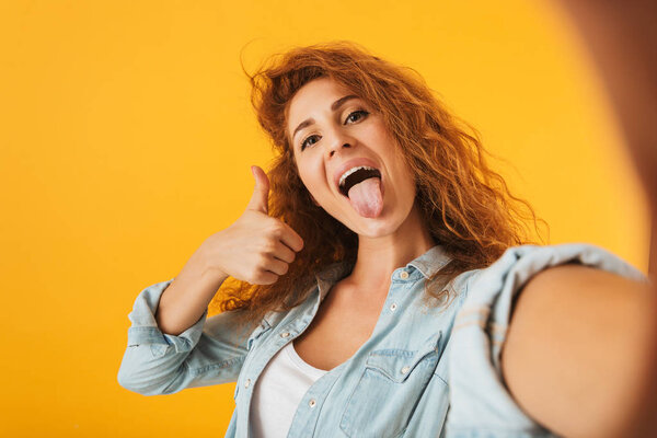 Image of funny joyous woman 20s showing thumb up with tongue sticking out while taking selfie photo isolated over yellow background