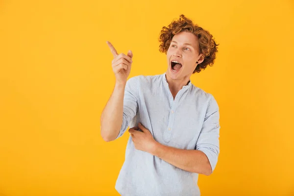 Portrait of astonished curly man 20s pointing finger and looking aside at copyspace with surprise isolated over yellow background