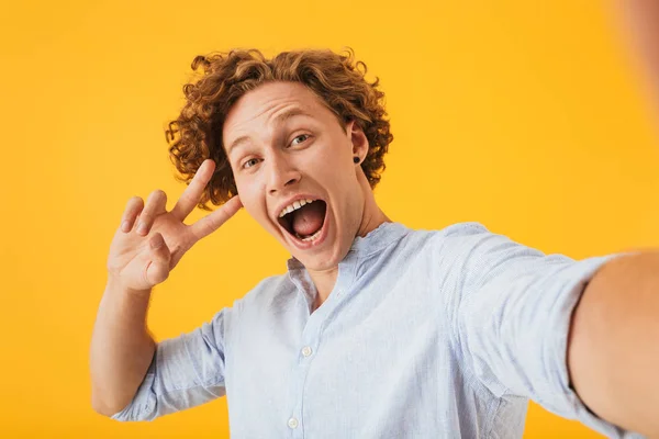 Portrait of ecstatic amazed man 20s taking selfie photo and showing peace sign isolated over yellow background