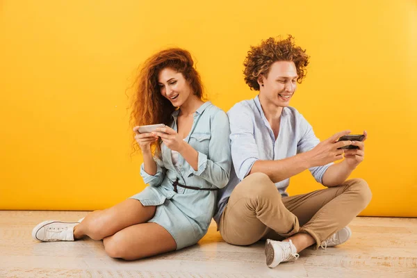 Photo of caucasian couple or friends attractive man and woman sitting on floor back to back and playing games on smartphones isolated over yellow background