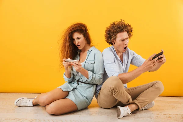 Image of excited couple or friends man and woman sitting on floor back to back and playing games on smartphones isolated over yellow background