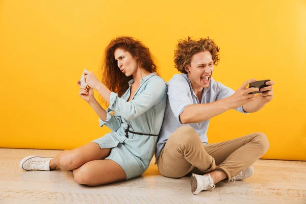 Portrait of joyful content couple or friends man and woman sitting on floor back to back and playing games on smartphones isolated over yellow background